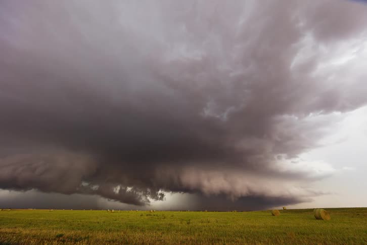 A wide low angle time lapse shows a vast dark shelf cloud slowly moving over a green field with round hay bales