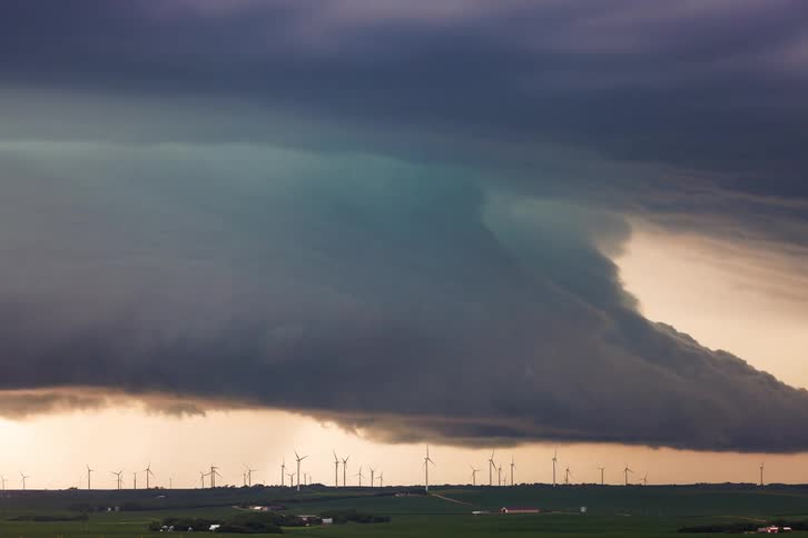 A wide view shows a large dark storm cloud with a rounded shelf like front moving across a sky lit by golden sunlight hovering low over a field with several wind turbines