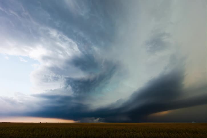 A large rotating supercell thunderstorm with a dark rounded base moves across a field under a swirling gray sky