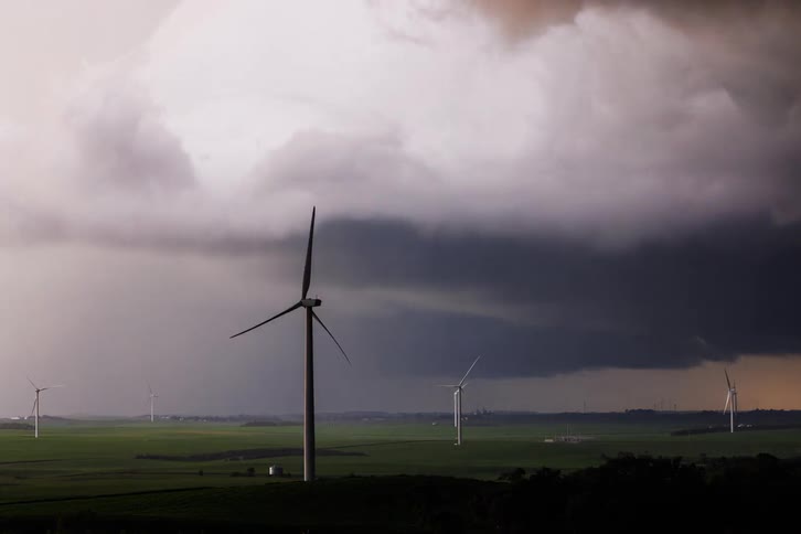 The video shows a dramatic shot of a wind farm under a stormy sky large white wind turbines stand in a green field as ominous dark storm clouds gather overhead in the distance flashes of lightning can be seen
