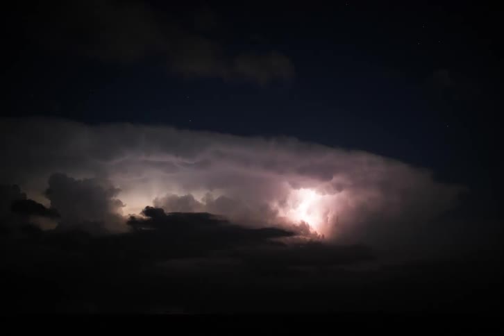 A series of lightning flashes illuminate a large storm cloud at night creating brief bright bursts of light behind the cloud