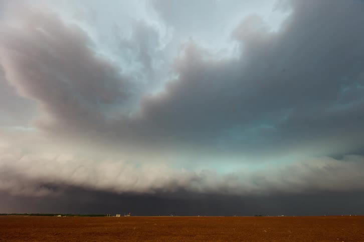 The video shows a timelapse of a massive and dark shelf cloud moving across a rural landscape the clouds flat smooth base looms over a wide flat reddishbrown field while the upper portion is characterized by a turbulent swirling mass of grey