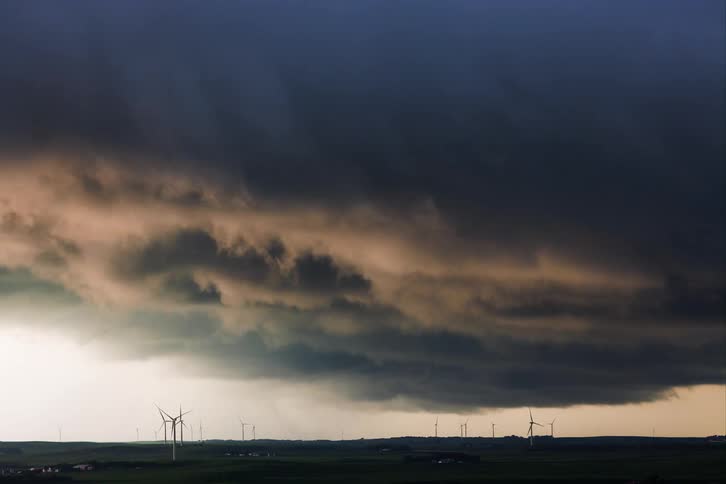 A massive shelflike storm cloud is moving ominously across a rural landscape dotted with wind turbines