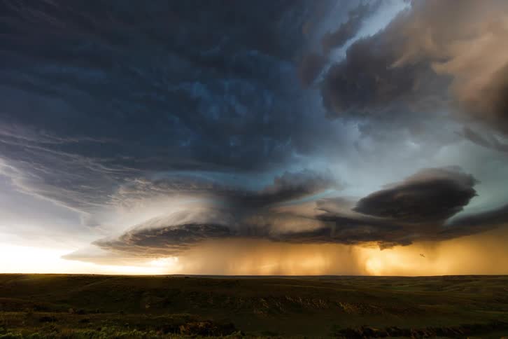 The video shows a spectacular timelapse of a massive and turbulent supercell thunderstorm gathering over a field at sunset as the sun sets a vast dark and swirling cloud structure forms and streaks of rain can be seen falling in the distance