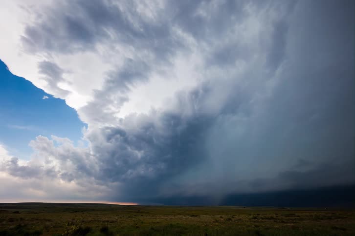 A massive dark supercell thunderstorm is dominating the sky contrasting with a small patch of blue sky and a distant horizon over a vast open field
