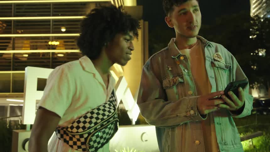 Two male friends consult a smartphone while moving around the city at night