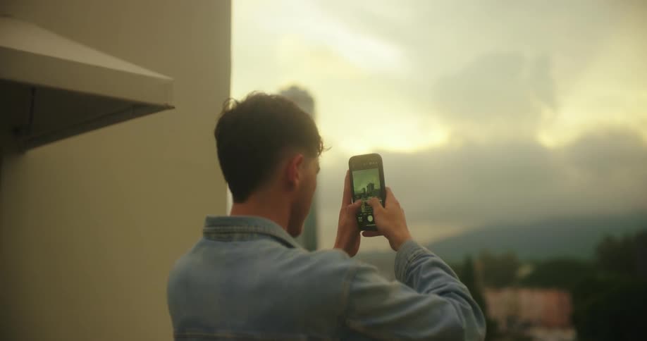 The video shows a man from behind standing on a balcony with a city in the background at sunset he holds his phone up to record a video capturing the view of a tall building and the sky