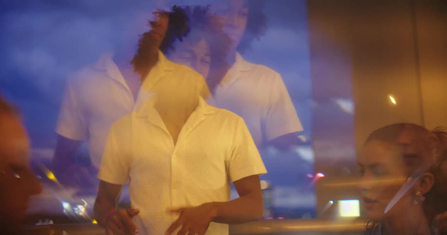 A man with dark curly hair and a white shortsleeved shirt is enthusiastically dancing while two woman are sitted having a conversation