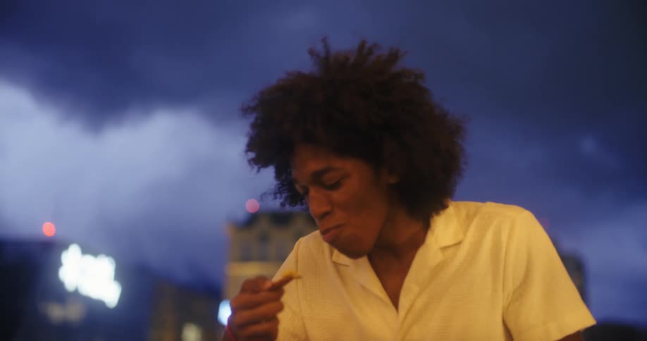A man with curly hair in a white shirt is eating something from his hand while another person is in the foreground