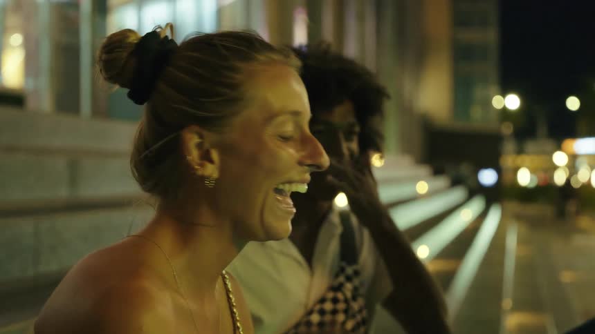 Two people a young woman and a young man sitting side by side on concrete steps on a city street while laughing and talking to someone else