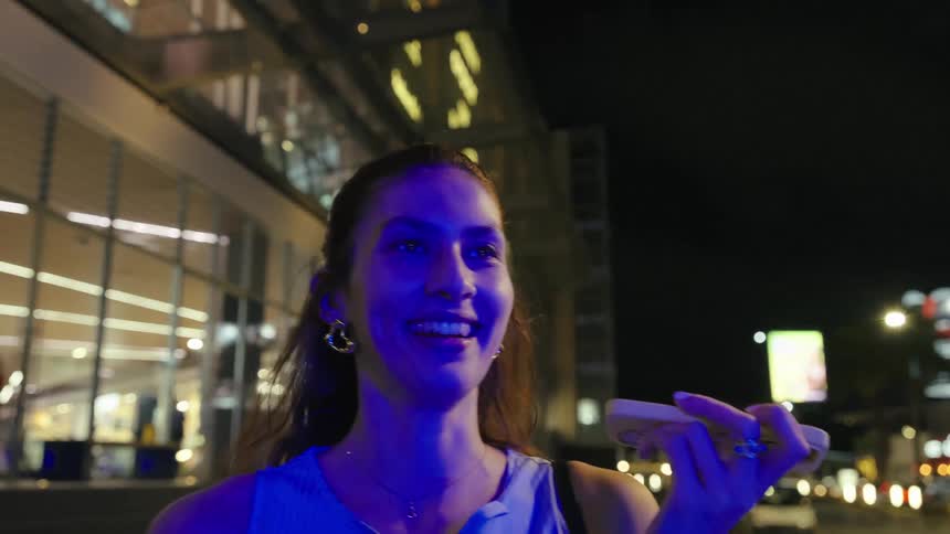 The video shows a young woman standing on a city sidewalk at night speaking into her phone using the speaker feature the background features a blurred street with a building and city lights
