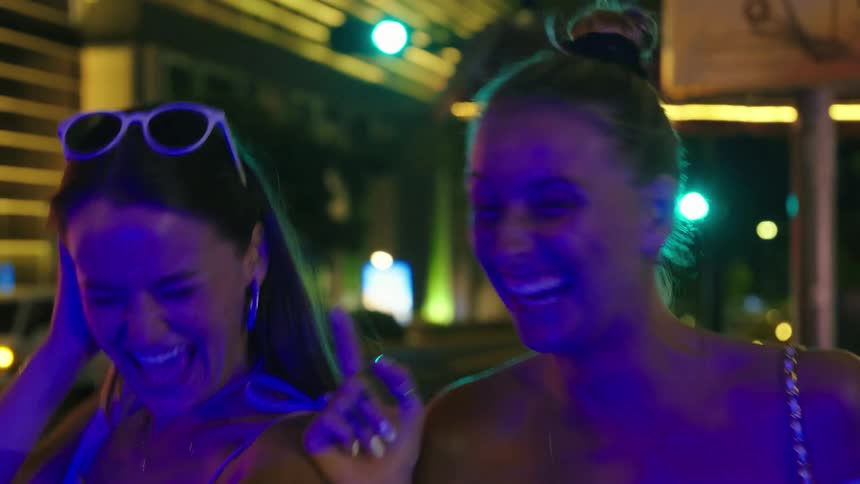 Two young women laughing and talking on a brightly lit street at night