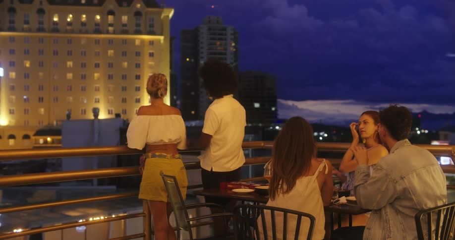 Friends talk while gathered together for an evening meal on a balcony overlooking the city