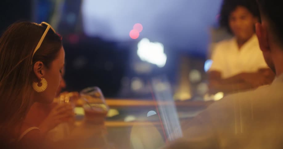 A group of friends drink cocktails at a rooftop bar after dark city lights turning into colorful bokeh beyond the glass railing