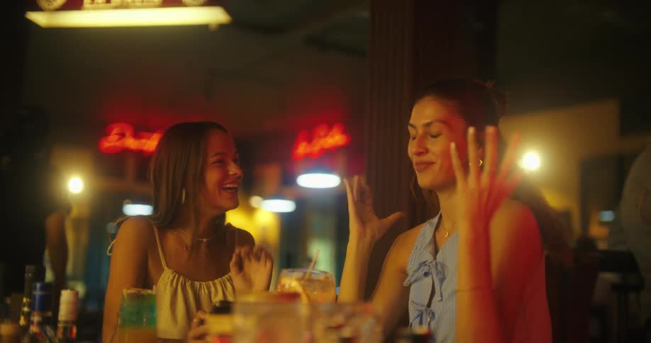 Two women smile while looking at something on a phone in a dimly lit bar