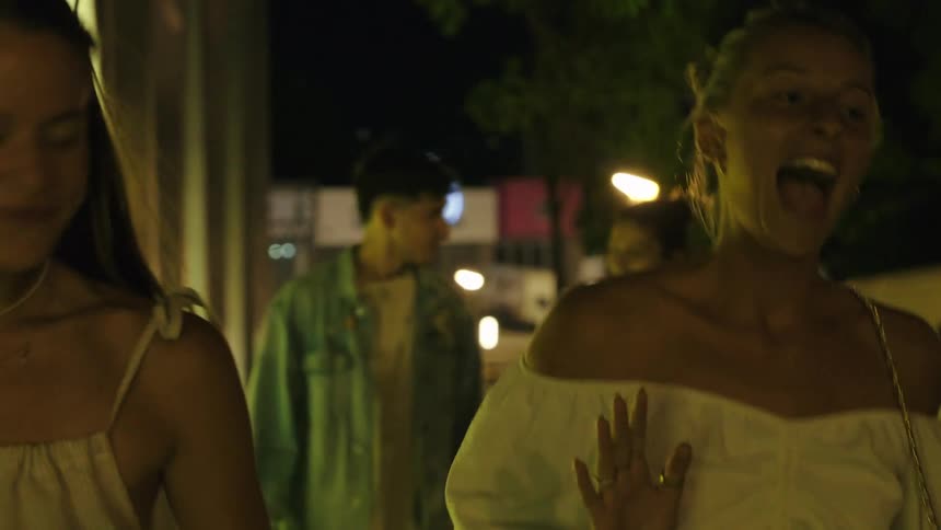The video shows a group of friends laughing and talking as they walk down a street at night the camera follows them before tilting down to focus on their feet and the pavement below as they continue walking