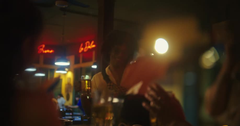 A video from a persons perspective shows a bartender standing behind a bar serving drinks to a group of friends who are blurred in the foreground