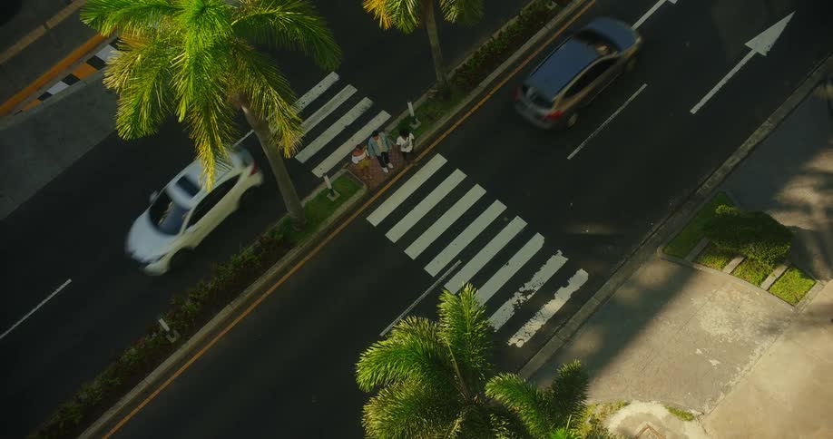 The video shows a highangle view of a crosswalk a small group of people waits at the curb while several cars drive past the people then start to cross the street