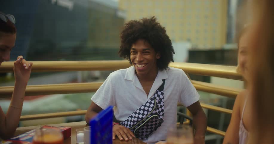 A young man with an afro smiles and gestures animatedly while sitting at a table on a balcony with another person with city buildings in the background