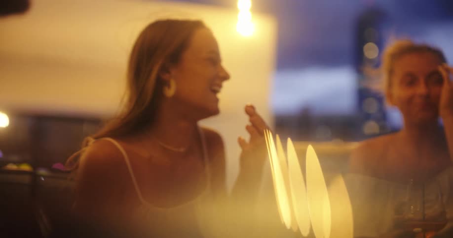 Two young women are enjoying drinks on a balcony at dusk with city lights and buildings visible in the background