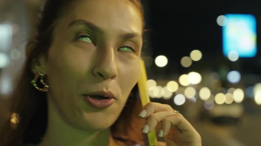 A young woman smiling as she talks on a cell phone on a city street at night while looking to the buildings around her
