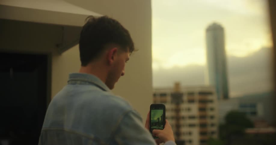 A young man standing on a building holds up is phone to take a photo