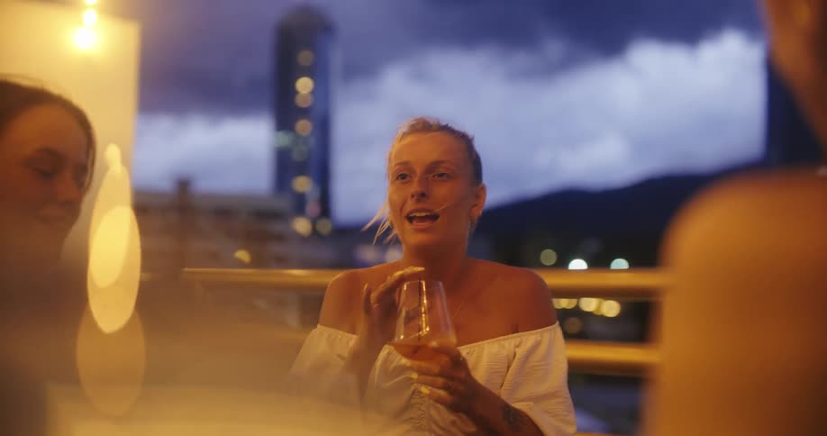 The video shows a group of young adults sitting at an outdoor table on a restaurant patio at night the woman in the foreground has her hair in a bun and is holding a glass while talking to her friends in the background there is a city and mountains lit