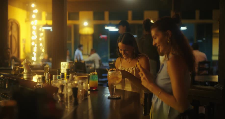 Two women laugh and take photos of their drinks at a lively bar