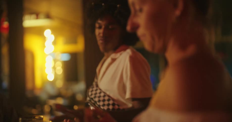 The video shows two men talking and laughing at a bar at night the man in the foreground is out of focus while the other man in the background is in focus the bar has warm lighting with several bright circular lights visible in the background the wat