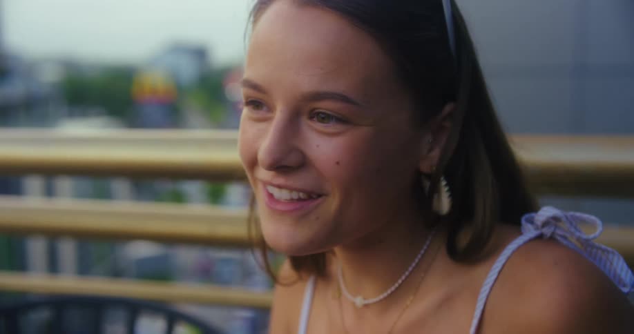 A woman sips a colorful beverage on a balcony at dusk while engaged in a conversation