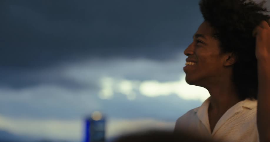 A young man with curly hair smiles as he holds up his phone to take a selfie with a blurred cityscape and cloudy sky in the background