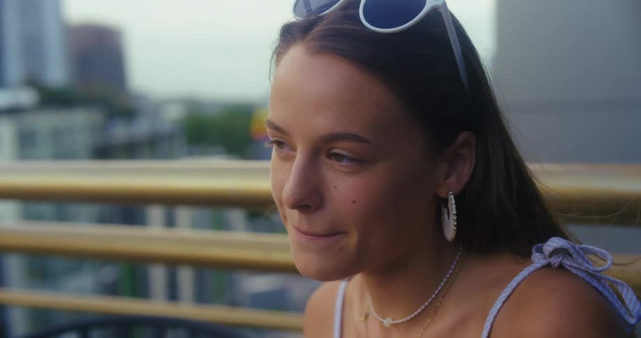 The video shows a young woman sitting outside drinking from a glass with a straw she takes a sip then smiles and laughs enjoying her beverage and the conversation with someone offcamera