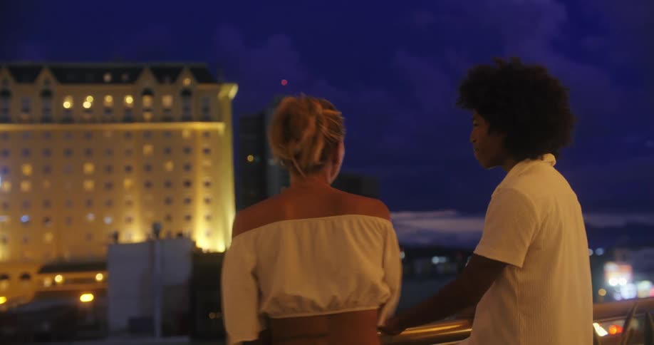 Black man and white woman standing on a balcony next to a rail laughing and talking with a metropolitan area on the background outdoors during night time