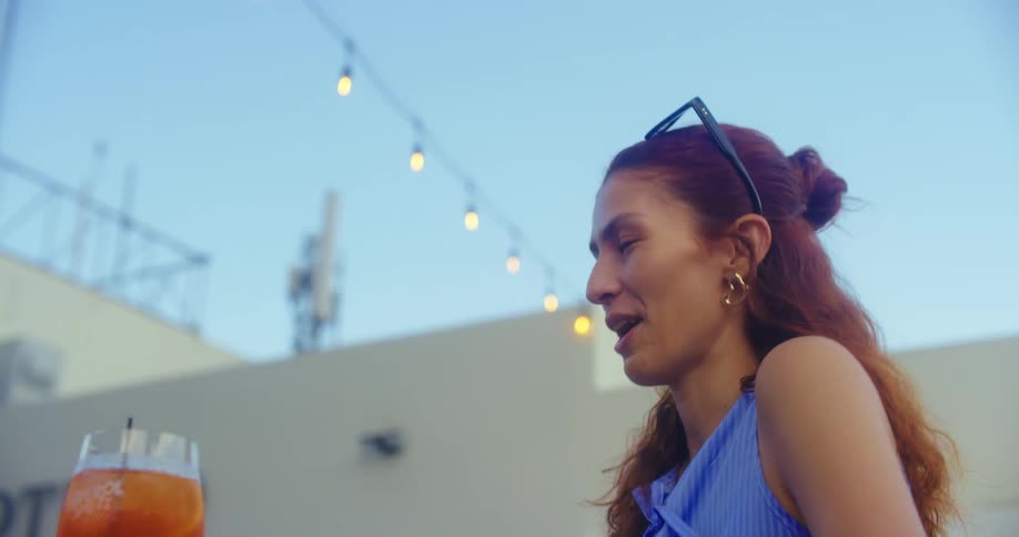 A woman with brown hair tied up in a bun wearing a light blue top and heart shaped earrings sits at a table with an orange drink and looks off screen while smiling and gesturing with her hands
