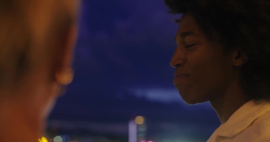 The video shows a side profile shot of a young man with dark curly hair smiling as he looks at someone offcamera the background is a blurry cityscape at night with colorful outoffocus lights