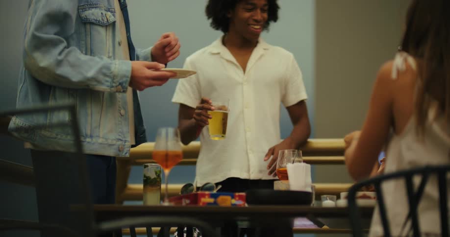 Friends are standing around a table with a man in a denim jacket and a man with dark curly hair exchanging a white plate and a third person with long brown hair sitting on the far right