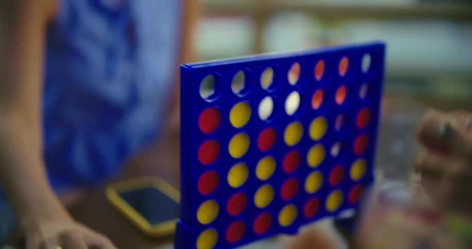 Two people are playing connect four with one persons hand dropping a yellow piece into the blue game board