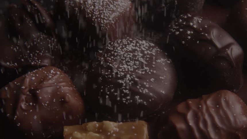 The video begins with a completely black screen and then fades in to show a closeup of a pile of various chocolates a fine white powder possibly powdered sugar is sprinkled over them creating a gentle snowfall effect