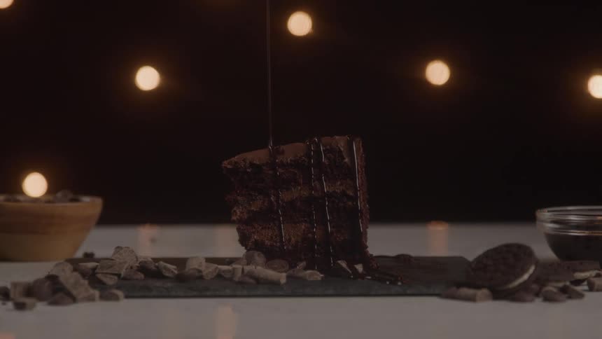 Melt chocolate drizzling over a chocolate cake surrounded by chocolate chips and a biscuit indoors