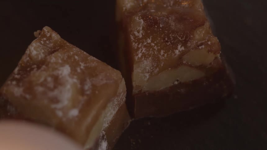 A closeup shot shows powdered sugar being dusted over two large rich pieces of homemade fudge that are sitting on a dark surface