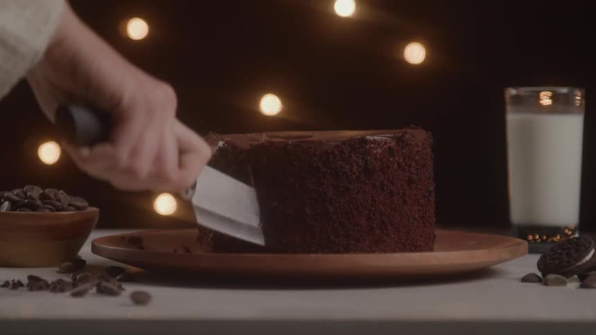 A womans arm using a knife to cut a slice from a chocolate cake in a bowl with a glass of milk beside it