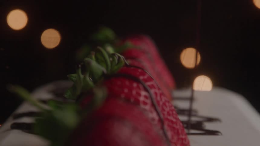 The video shows a closeup shot of a row of fresh strawberries being drizzled with a thick chocolate sauce the focus is on the rich red of the berries and the dark glossy chocolate against a background of soft outoffocus lights