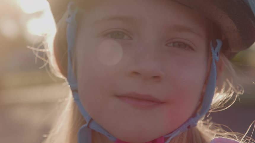 The video shows a closeup shot of a young girl with blonde hair wearing a bike helmet she is smiling and looking at the camera with the bright sun flaring in the background