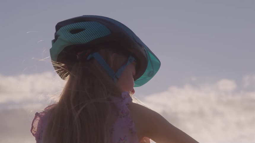 Young blonde girl wearing a pink dress and a helmet riding a bike outdoors during sunset