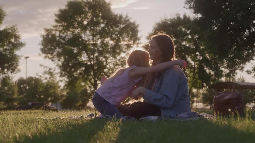A mother and daughter sit on a blanket in a grassy park with the mother gently touching the daughters face before they embrace