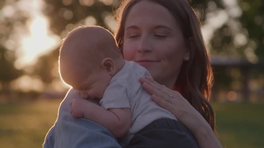 Young white woman holding a sleeping baby in her arms and tapping his back gently while smiling surrounded by trees with the sun setting on the background outdoors