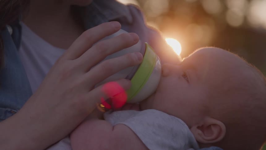 Baby drinking milk from a baby bottle held by female white hands outdoors during dusk