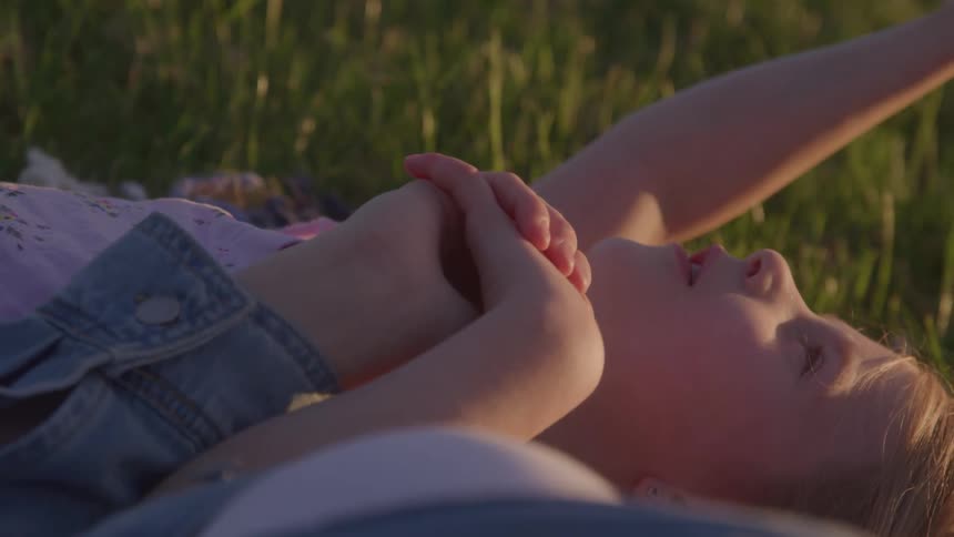 A child reclines in tall grass at golden hour clasping their hands and gazing upward with a soft thoughtful expression