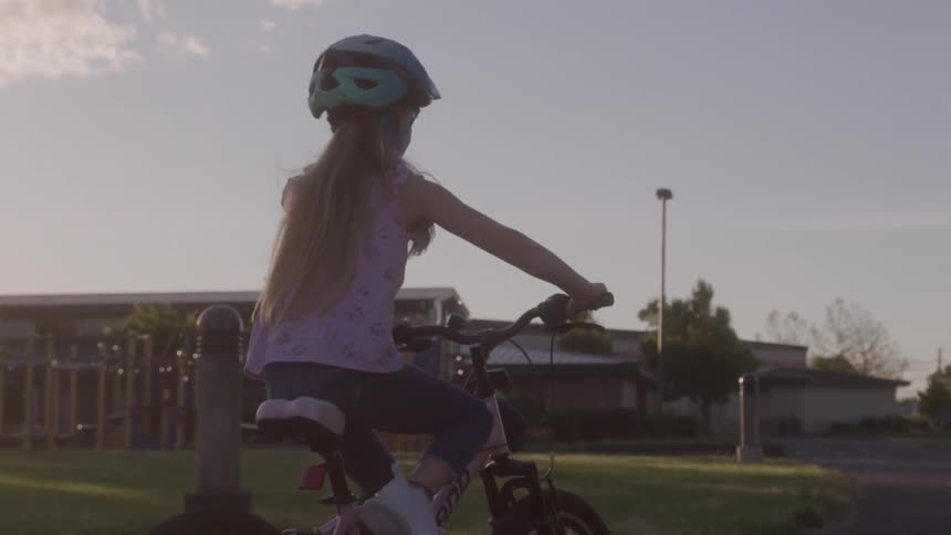 A child with long blonde hair and a teal helmet rides a pink bicycle toward a school building and a playground as the sun sets