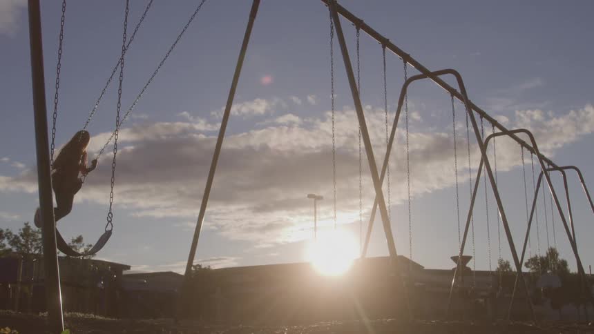 A young girl with long hair swings on a swing set at a park during sunset silhouetted against the bright sky